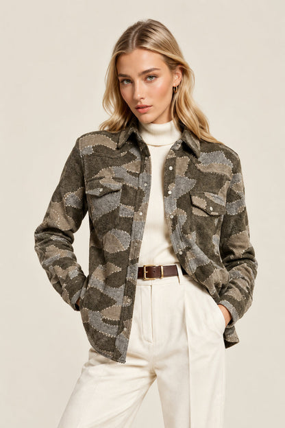 A woman modeling a cropped button-up shirt jacket with a green, brown, and gray camouflage pattern, worn over a cream turtleneck and cream wide-leg trousers.